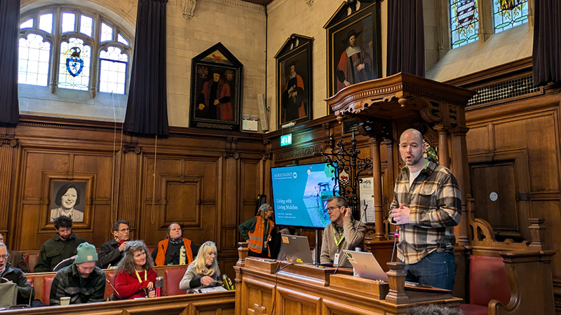 Presenters and audience in a panelled room with paintings and stained glass windows